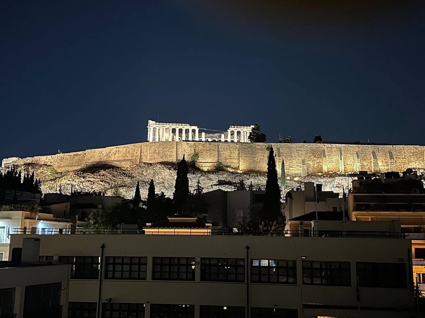 The floodlit Acropolis and Parthenon seen at night from the rooftop terrace of the building, above the neighbouring Mets rooftops